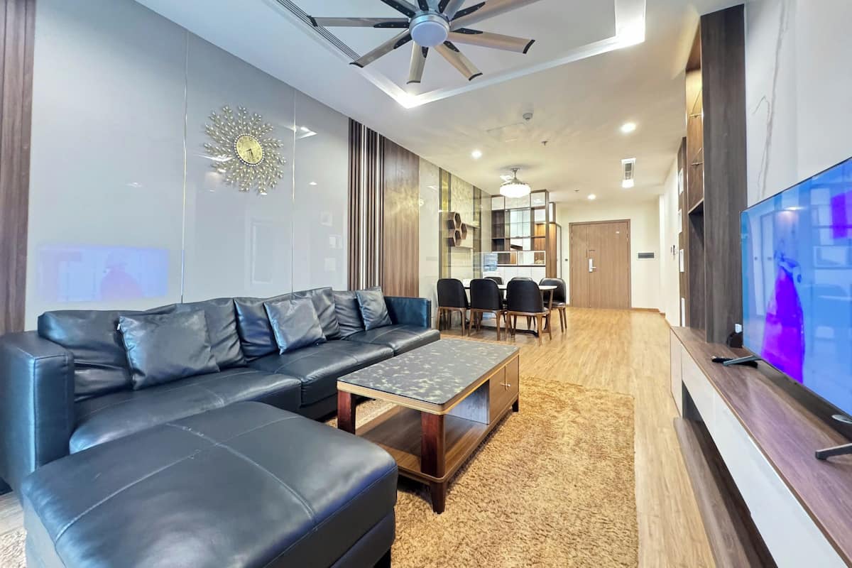 A modern living room with a sectional sofa, a coffee table, a dining area with a table and chairs, a large flat-screen TV, and a ceiling fan.