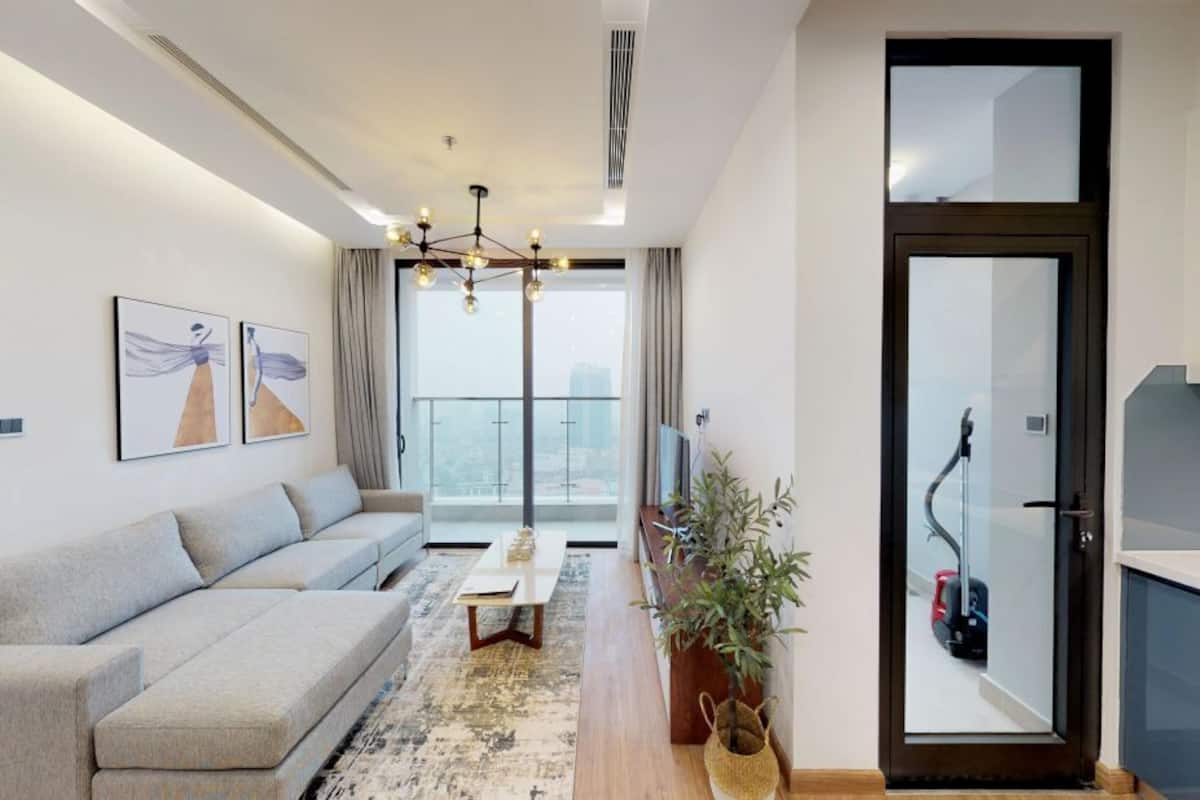 A modern living room with a sofa, a coffee table, and a potted plant. There is a door leading to a balcony with a view of the city.