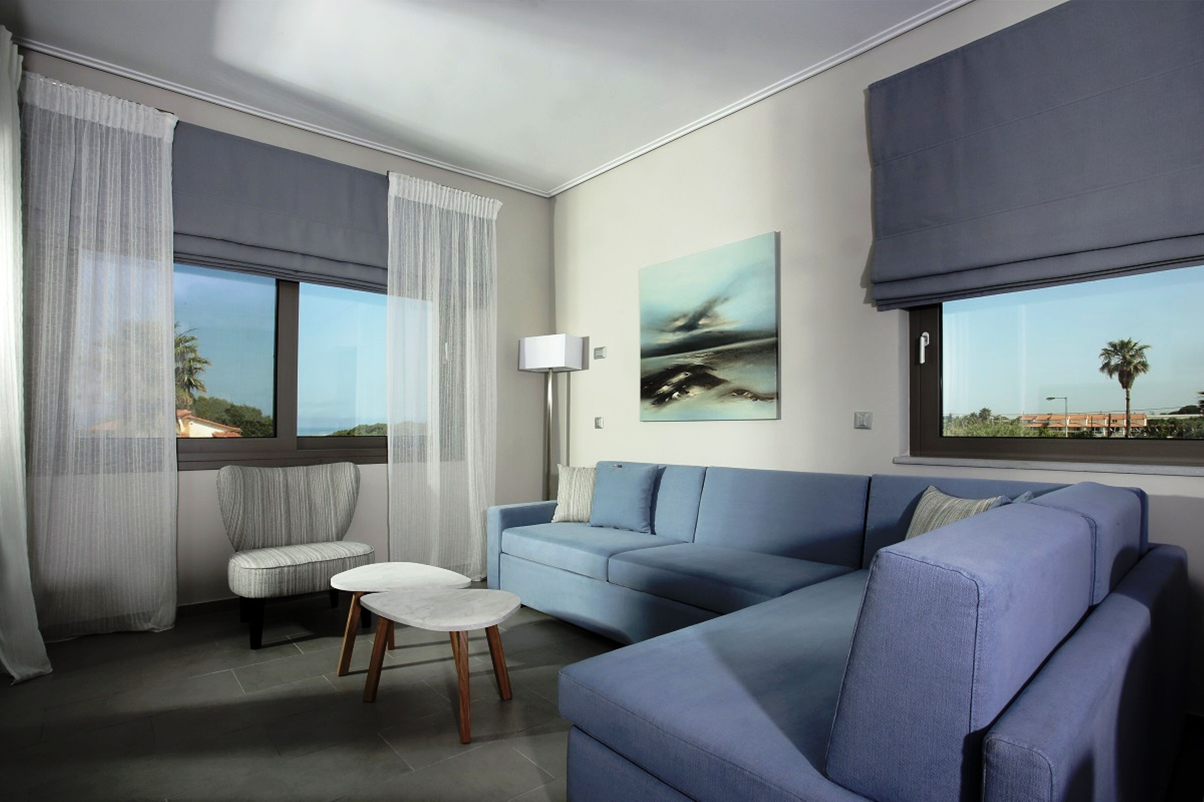 A modern living room with a blue sofa, a small round table, and a chair. There is a large window with sheer curtains, a painting on the wall, and a lamp on a side table.