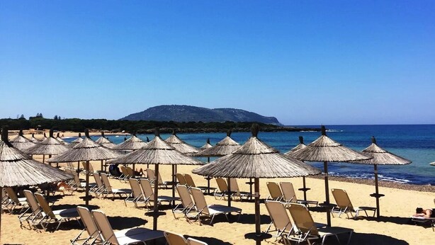 A beach with rows of sun loungers and thatched umbrellas, a clear blue sky, and a mountain in the background. - Hotel Lagouvardos Village (Trifylia)
