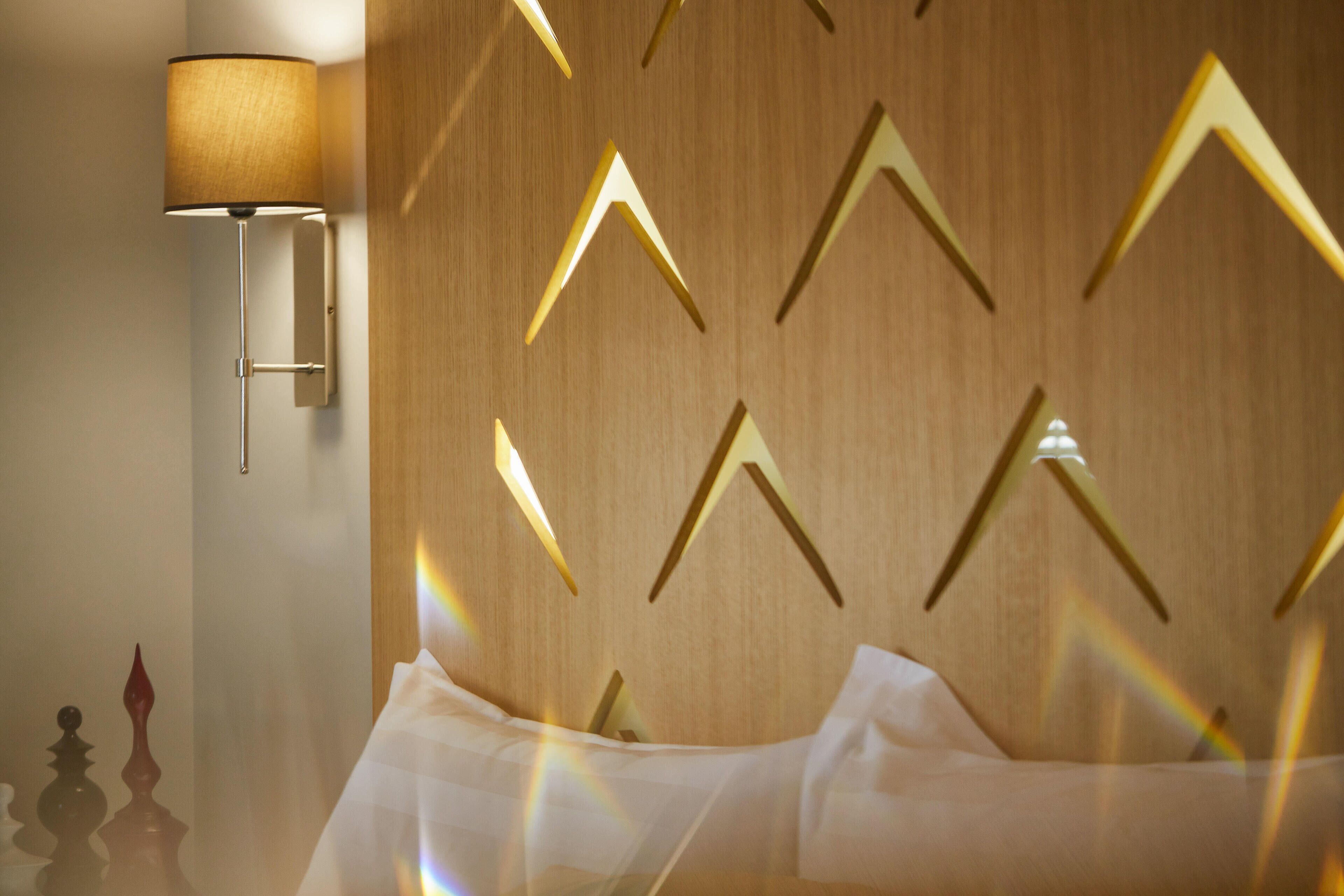 A hotel room with a bed, a wall-mounted lamp, and a wooden wall with geometric patterns.