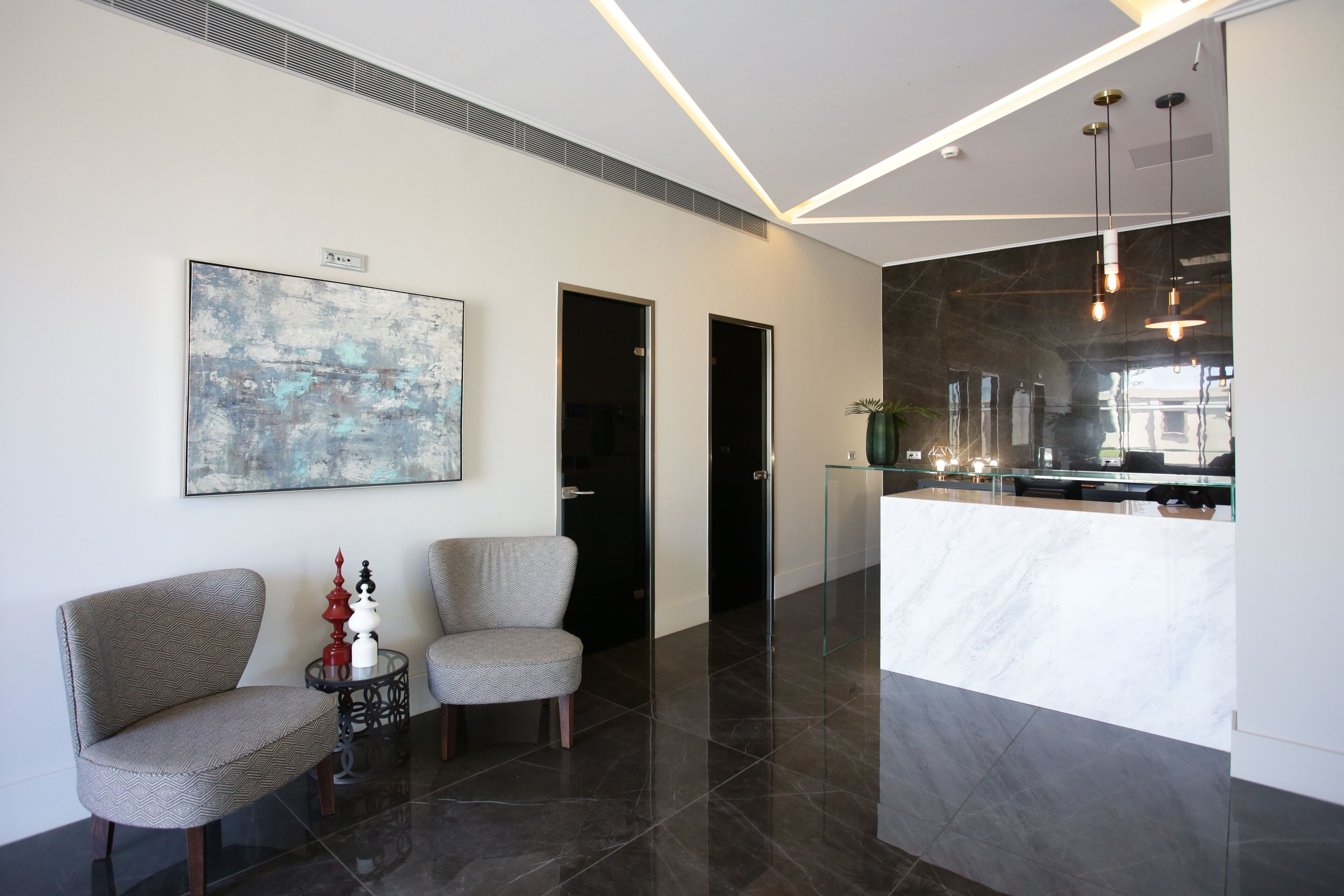 Eine moderne Lobby mit zwei gepolsterten Sesseln, einem Glastisch mit Dekoration, einem großen abstrakten Gemälde und einer Rezeption mit Marmorplatte.