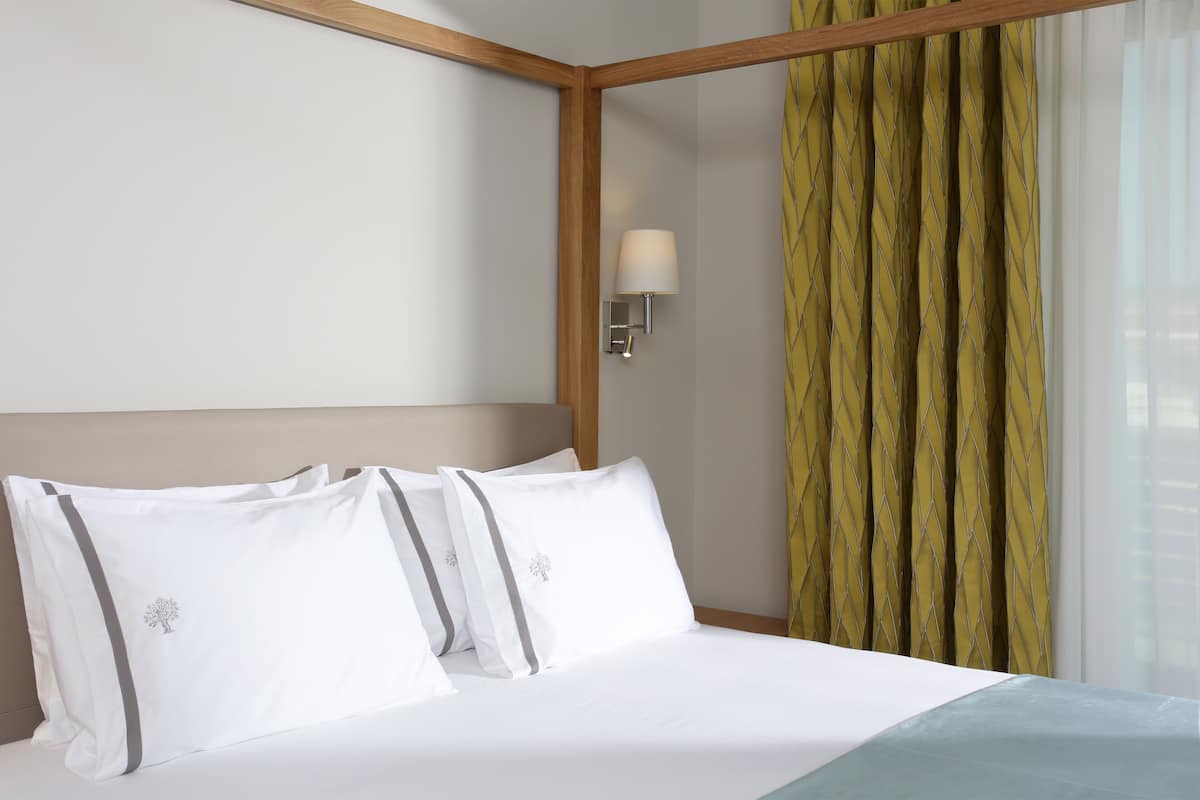 Ein Hotelzimmer mit Bett, weißer Bettwäsche, einem Nachttisch mit Lampe und einem Fenster mit Vorhängen.