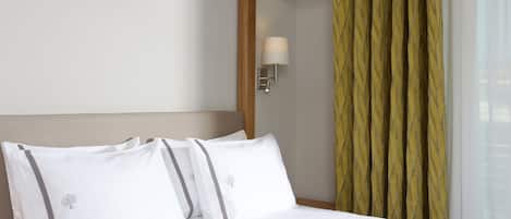 A hotel room with a bed, white linens, a nightstand with a lamp, and a window with curtains.