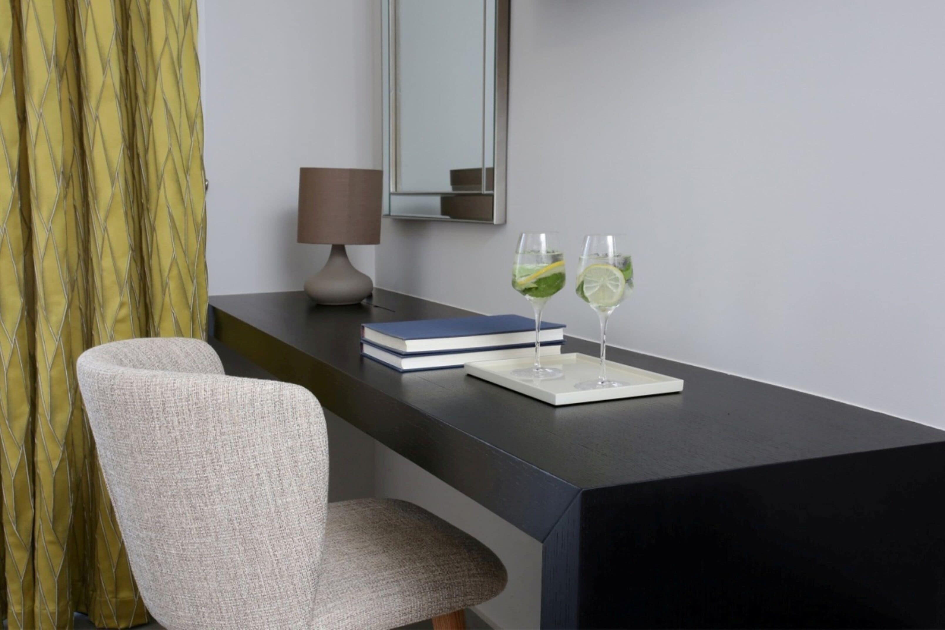 A modern room with a dark desk, a chair, a lamp, a mirror, and two glasses with lime slices on a tray.