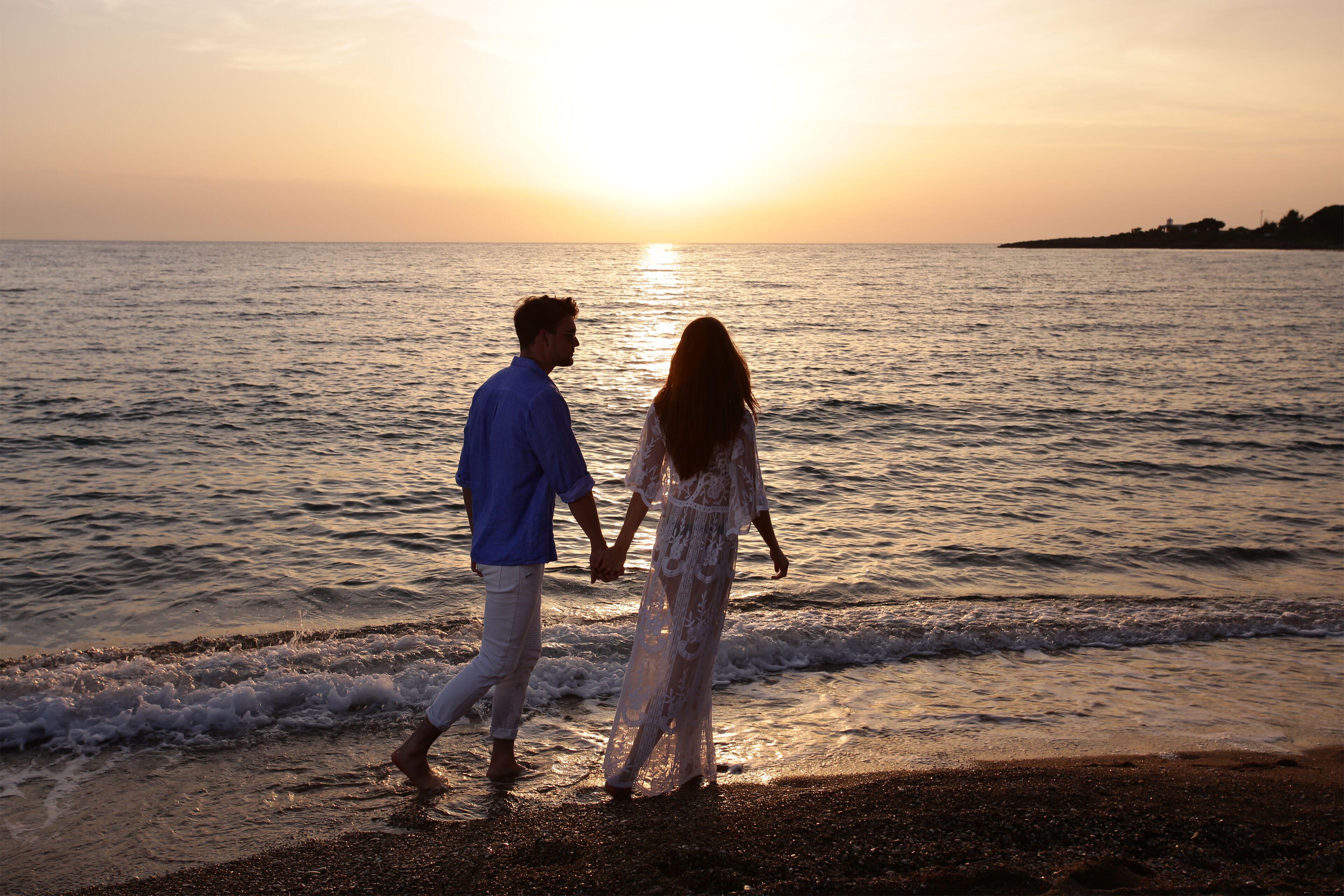A couple walking hand in hand on a beach at sunset.