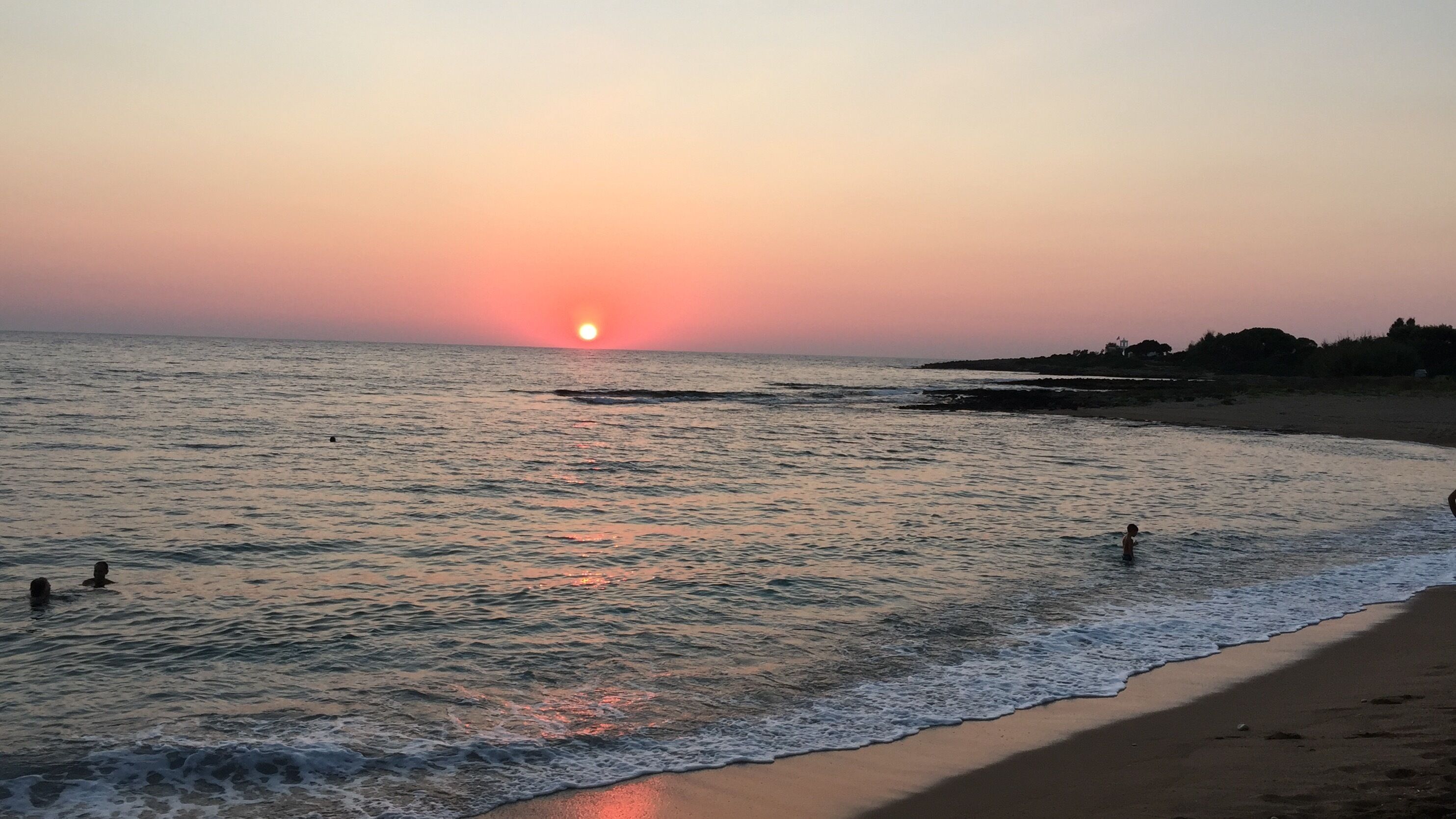Una playa con el sol poniéndose sobre el océano, aguas tranquilas y algunas personas nadando.