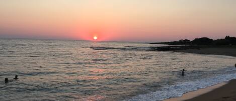 Una playa con el sol poniéndose sobre el océano, aguas tranquilas y algunas personas nadando.