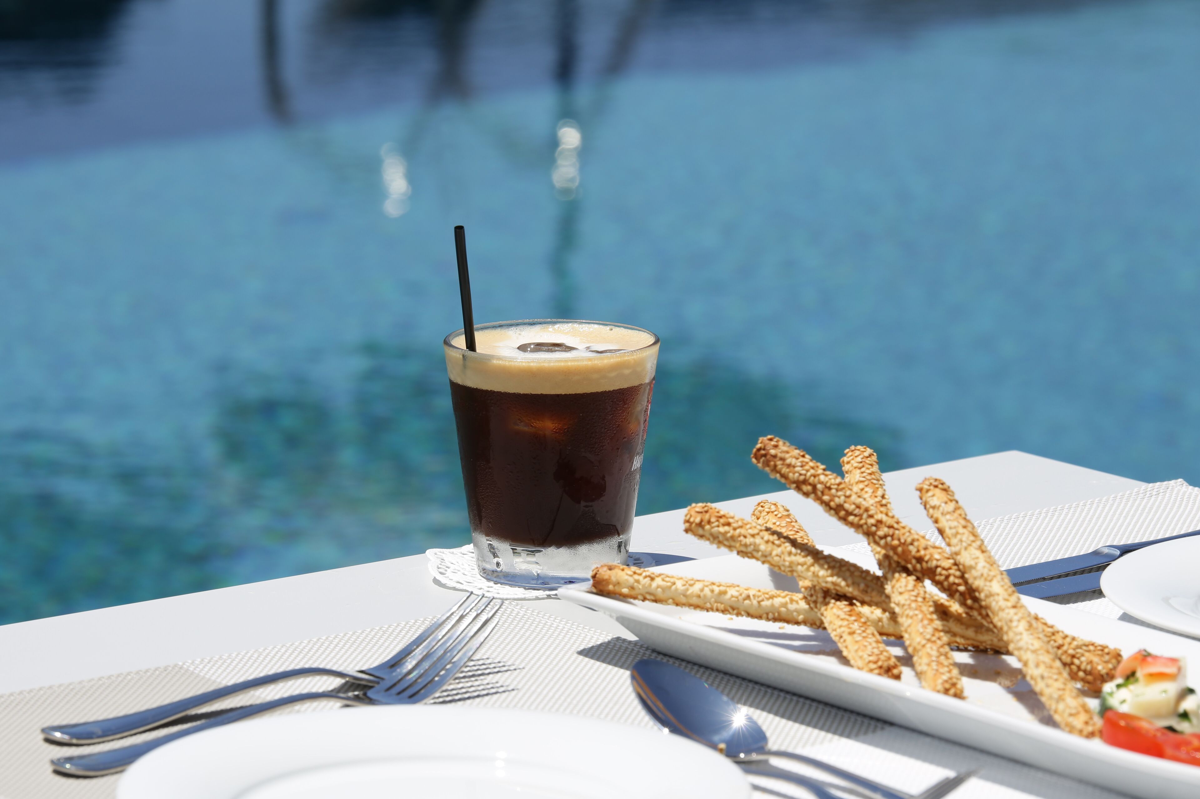 Un vaso de café helado con pajita, un plato de aperitivos crujientes, y un tenedor y cuchara sobre una mesa junto a una piscina.