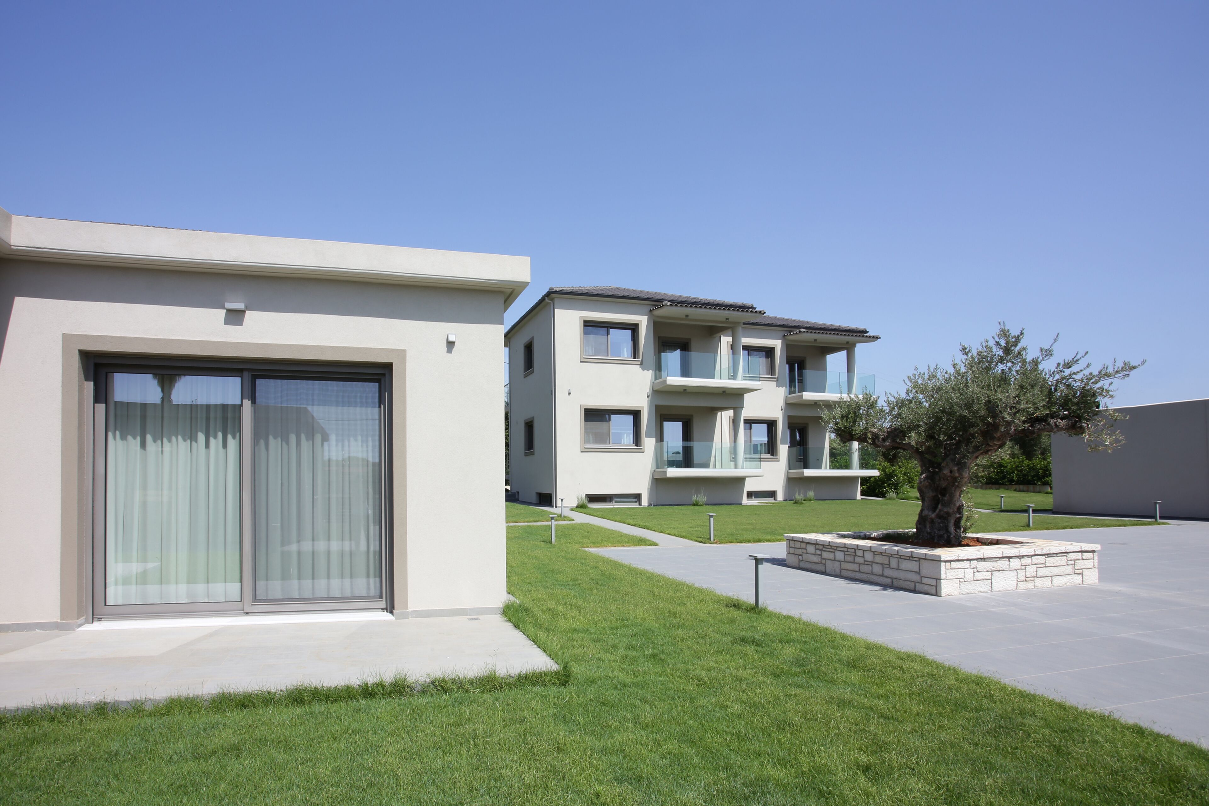 A modern residential building with large sliding glass doors, a well-maintained lawn, and a small tree in a planter.
