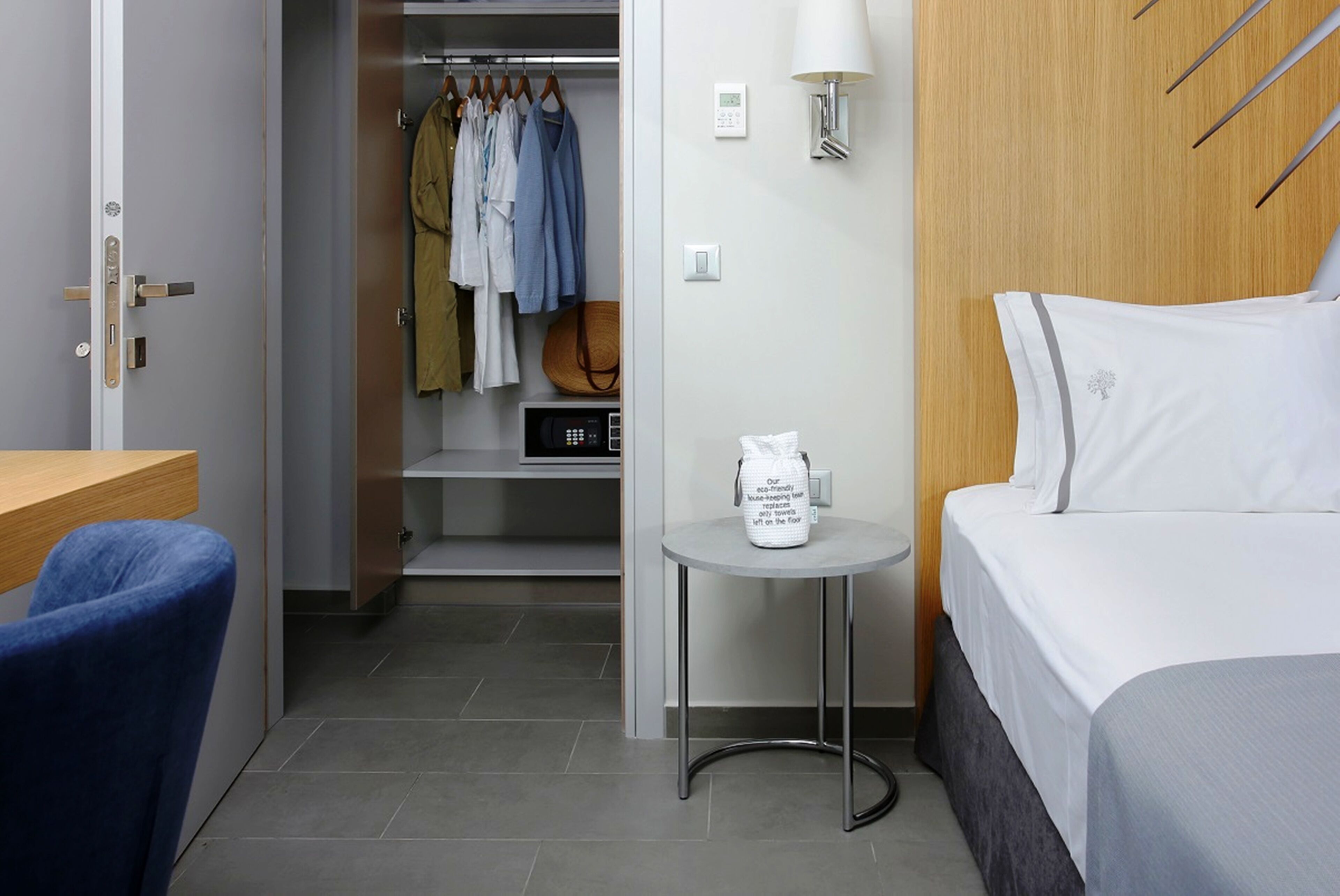 Une chambre d’hôtel comprenant un lit, un bureau, une chaise, une armoire avec des vêtements suspendus, une petite table avec un sac, et une lampe fixée au mur.