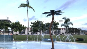 Ein öffentlicher Brunnen mit mehreren Wasserstrahlen, umgeben von Palmen und einer Rasenfläche.