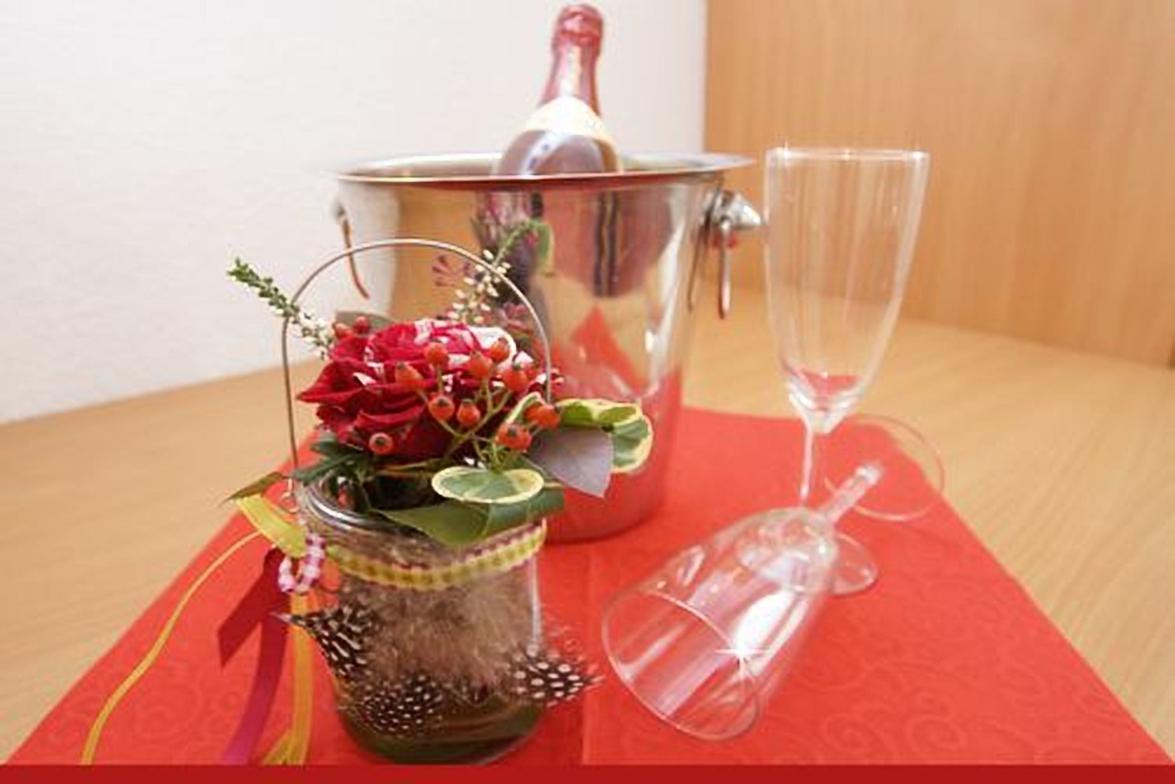 Uma mesa com toalha vermelha, um copo de vidro transparente, uma panela de metal e um frasco decorativo com bago e folhas verdes.