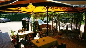 An outdoor seating area with tables and chairs under a yellow umbrella, surrounded by potted plants and a wooden fence. - Hotel & Restaurant zur Linde (Leuna)