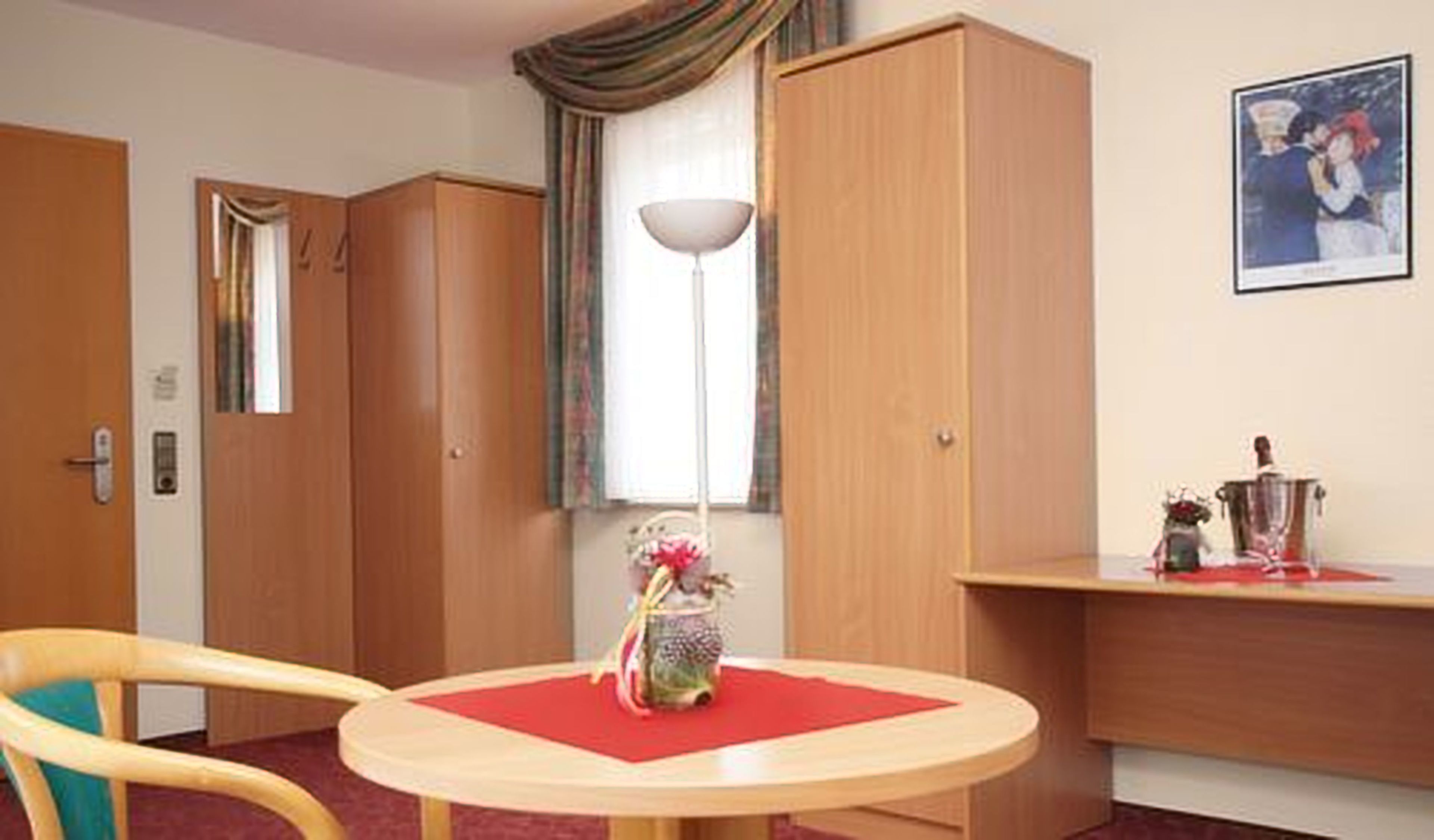 A hotel room with a round table, a chair, a lamp, a wardrobe, and a picture on the wall.