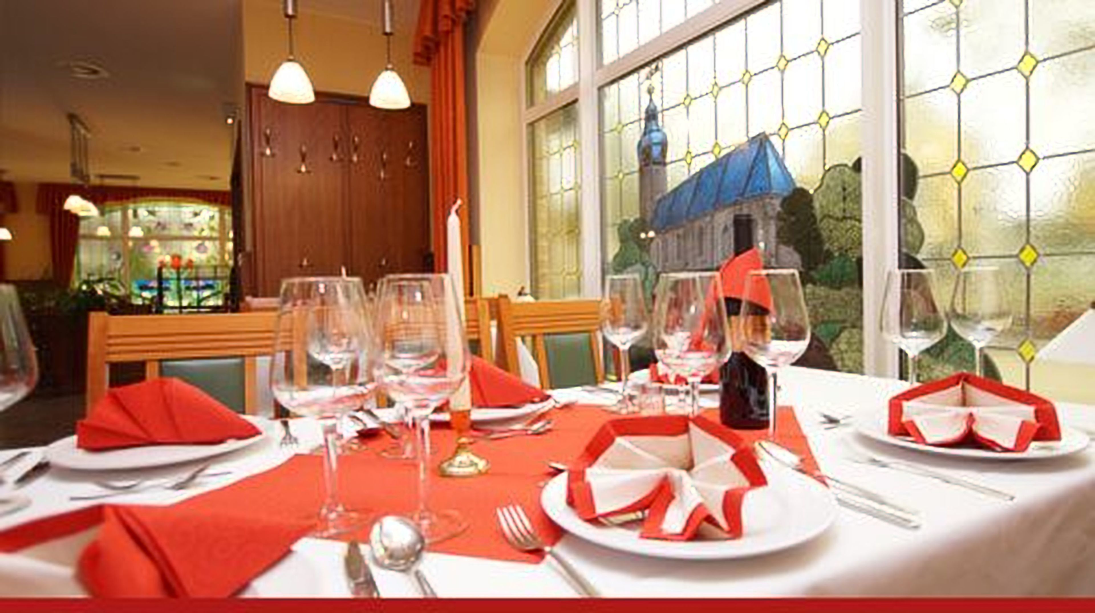 Una mesa de comedor dispuesta con servilletas rojas, copas de vino y platos, con un vitral de fondo.