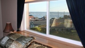 Een kamer met een groot raam dat uitkijkt op een kustgebied, een bed met beddengoed met een patroon, en een lamp op een nachtkastje.