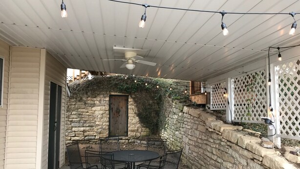 An outdoor patio with a stone wall, a round table with chairs, string lights, and a ceiling fan. - A perfect weekend gateway! (Grafton)