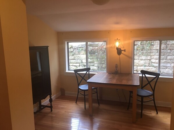 Un área de comedor con una mesa y sillas de madera, una lámpara fijada a la pared y vista a una pared de piedra a través de una ventana.