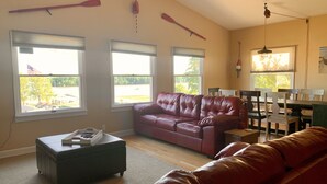 A spacious living room with a leather couch, a coffee table, and a dining area with wooden chairs and a table. Large windows provide a view of the outdoors. - A perfect weekend gateway! (Grafton)