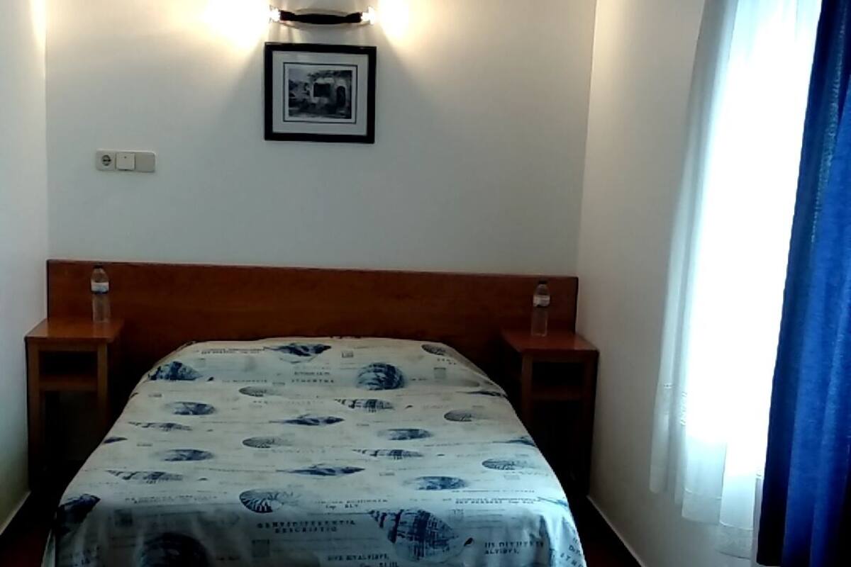 A bedroom with a bed, two bedside tables, a wall-mounted light, a framed picture, and a window with curtains.