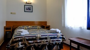 Una cama bien hecha con una colcha a rayas, dos mesitas de noche, un banco pequeño de madera, una lámpara fijada a la pared e imagen en un marco colgado.