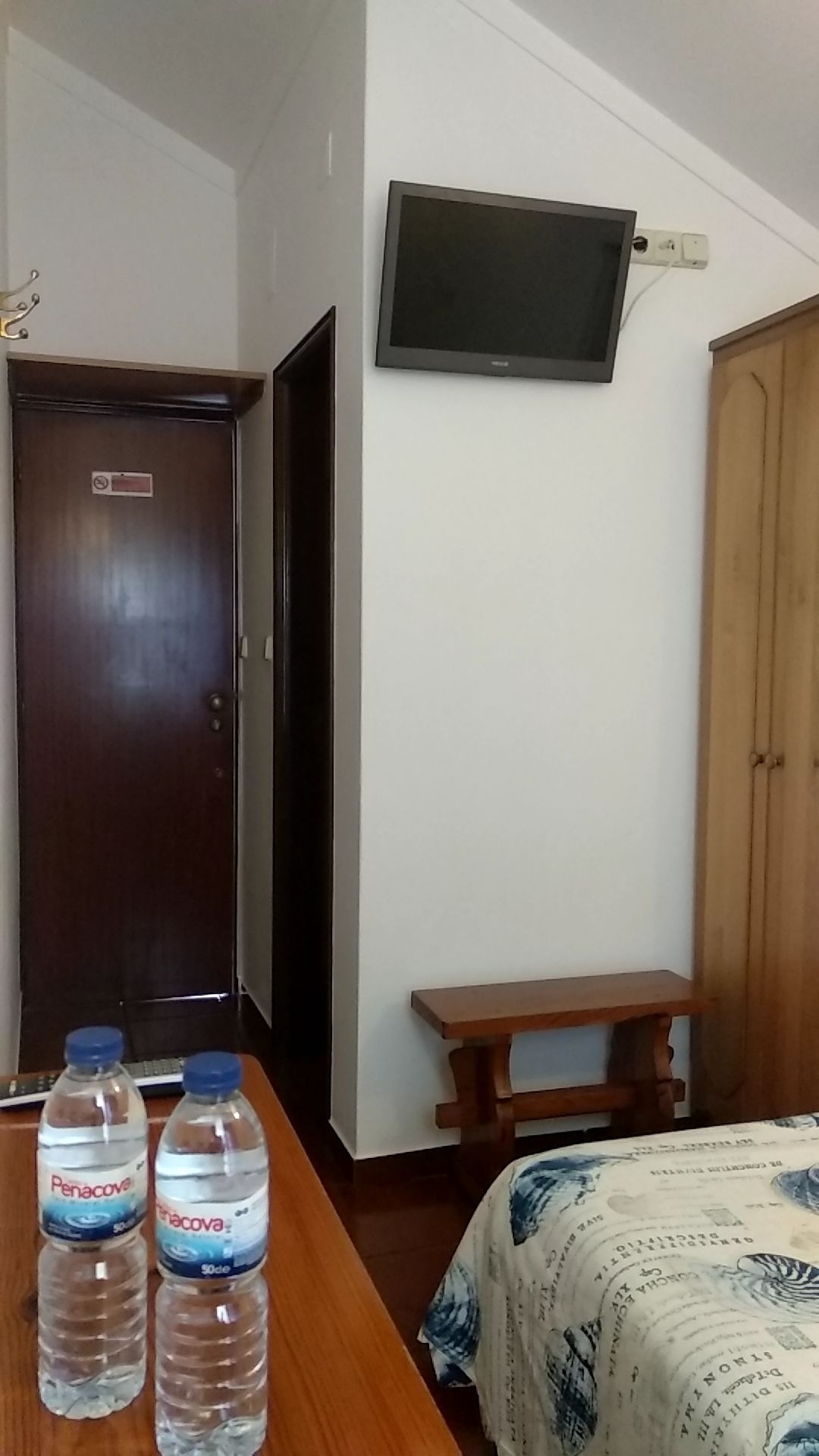Een hotelkamer met een bed, een bureau met twee waterflesjes, een aan de muur bevestigde televisie en een houten garderobe.