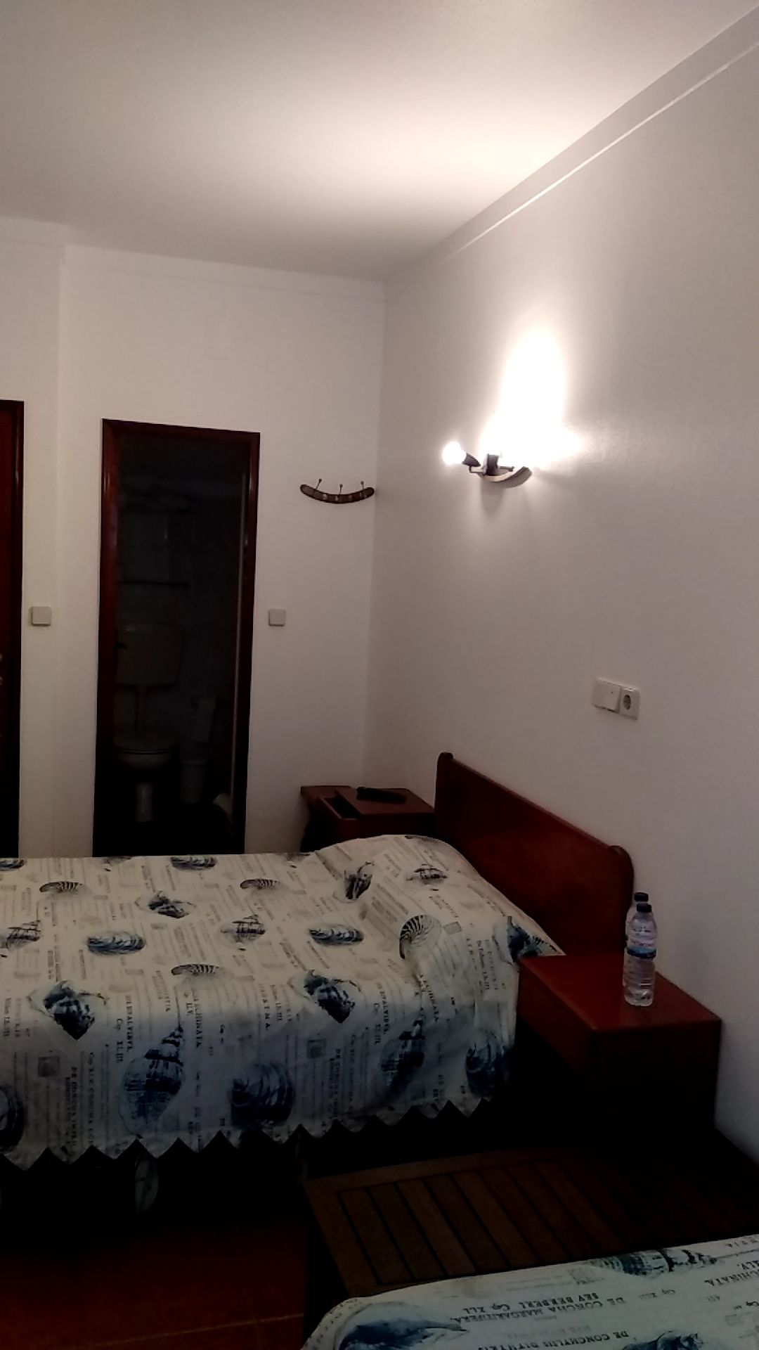 A small, simple room with a bed, a nightstand, a wall-mounted light, and a door.