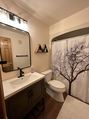 A bathroom with a vanity, a toilet, a shower curtain with a tree design, and a mirror with lights.