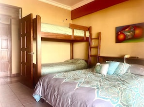 A room with a bunk bed, a door, a bed with patterned bedding, and a wall with a fruit painting. - Million Dollar View in Heart of Flamingo w/Costa Rican Decor! (Guanacaste)