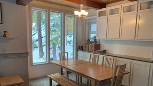 Una cucina con tavolo e sedie in legno, mobili bianchi e ampie finestre con vista sugli alberi.
