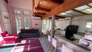 Cucina moderna con frigorifero nero, microonde e macchina del caffè. Zona pranzo con una sedia rossa e un divano grigio. La stanza ha un soffitto in legno e ampie finestre.