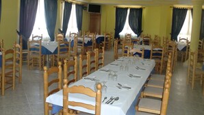 Un comedor con varias mesas dispuestas para una comida, sillas de madera y cortinas azules.