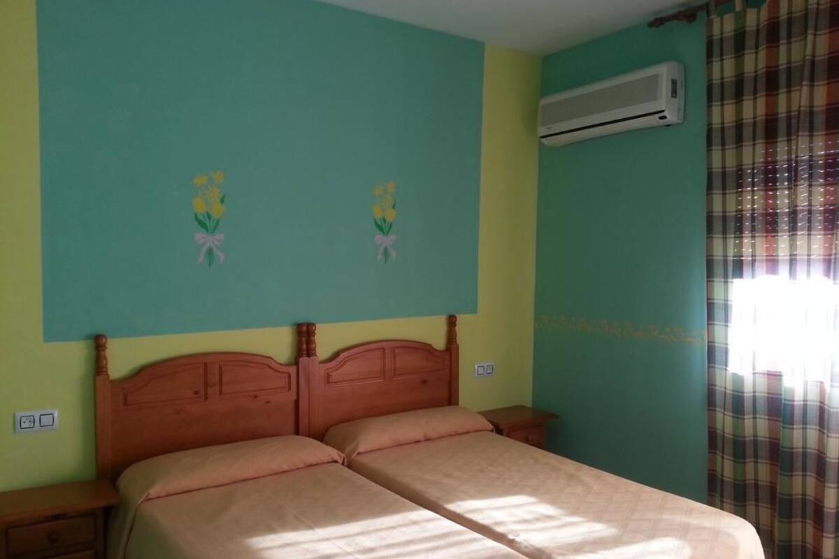 Un dormitorio con una cama doble, dos mesitas de noche, aire acondicionado de pared y una ventana con cortina escocesa.