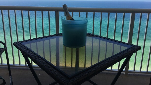 Balcón con una mesa de vidrio y un cubo azul que contiene una botella, con vista a un océano azul y despejado.