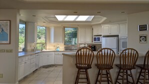 A spacious kitchen with white cabinetry, a central island with bar stools, a skylight, and a view of greenery outside. - Spacious 5-bedrm home:Ocean Views,hot tub,BBQ,pool & ping pong tables, fast wifi (Bodega Bay)
