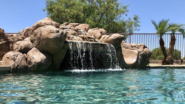 A clear pool with a waterfall feature surrounded by large rocks and palm trees. - Resort-Style Oasis | Pool & Spa | Waterslide | Basketball Court (Gilbert)
