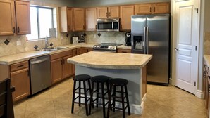 A modern kitchen with wooden cabinets, stainless steel appliances, a large island with stools, and a window providing natural light. - Resort-Style Oasis | Pool & Spa | Waterslide | Basketball Court (Gilbert)