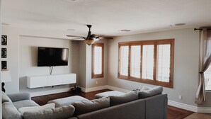 A living room with a grey sectional sofa, a flat-screen TV, a ceiling fan, and a window with wooden shutters. - Resort-Style Oasis | Pool & Spa | Waterslide | Basketball Court (Gilbert)