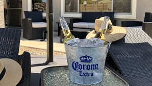 A patio with outdoor furniture, a glass table, and a bucket labeled "Corona Extra" containing bottles. - Resort-Style Oasis | Pool & Spa | Waterslide | Basketball Court (Gilbert)