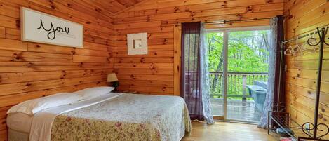 Un dormitorio de cabaña de madera con cama, mesita de noche, ventana con cortinas y vistas al exterior.