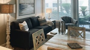 A living room with a dark-colored sofa, a wooden coffee table, and a dining table with chairs. There is a ceiling fan, a painting on the wall, and a window with blinds. - Prime October Dates Are Open! Perfect Time For a Beach Trip! (Orange Beach)