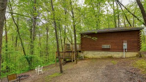 A rustic cabin with a wooden deck and a brick foundation, surrounded by a forested area with benches. - Blue Rose Cabins - Pine Ridge Cabin (Logan)