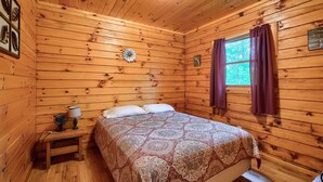 A wooden cabin room with a bed, a nightstand, a lamp, and a window with curtains. - Blue Rose Cabins - Pine Ridge Cabin (Logan)
