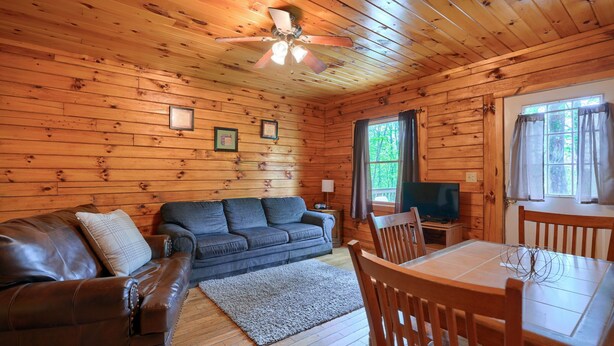 A cozy cabin interior with a wooden ceiling, a sofa, a dining table with chairs, and a television. - Blue Rose Cabins - Pine Ridge Cabin (Logan)