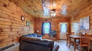 A wooden cabin interior with a leather sofa, a dining table with chairs, a ceiling fan, and a television. - Blue Rose Cabins - Pine Ridge Cabin (Logan)
