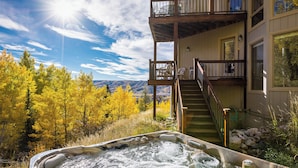 A hot tub with a view of autumn trees and mountains, a wooden deck, and a staircase leading to a balcony.