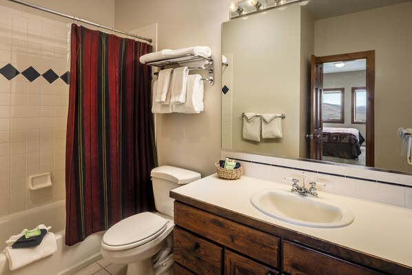 A bathroom with a white toilet, a shower with a striped curtain, a sink with a mirror, and a towel rack with white towels. - An Easy 3-minute, 150 Yard Walk to the Slopes! (Steamboat Spring)