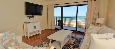 A living room with a flat-screen TV, a white sofa, a coffee table, and a view of the beach through a sliding glass door.