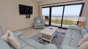 Una sala de estar con televisor grande, un sofá, un sillón y vista a la playa.